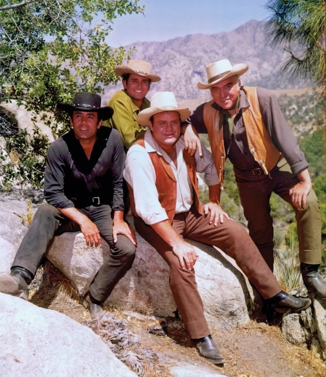 The Cartwright family of Bonanza gathered outdoors in a scenic location. From left to right: Pernell Roberts as Adam, Michael Landon as Little Joe, Dan Blocker as Hoss, and Lorne Greene as Ben Cartwright