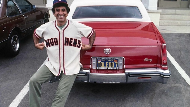 Jamie Farr posing in a Toledo Mud Hens jersey next to his car with a personalized Toledo license plate, showcasing his real-life connection to his hometown and love for baseball