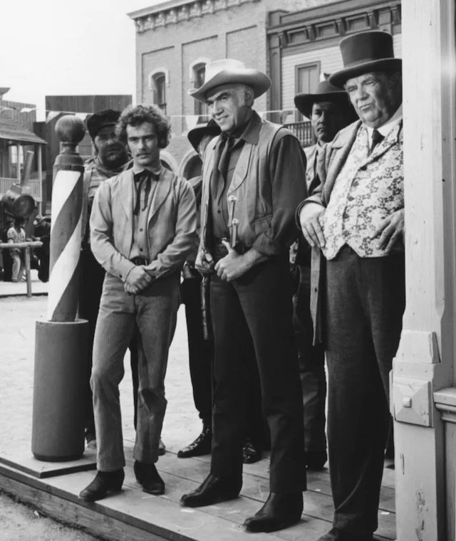 A scene set in town from Bonanza, with Ben Cartwright standing among other men. The group looks on with serious expressions, likely witnessing or discussing an important event in town