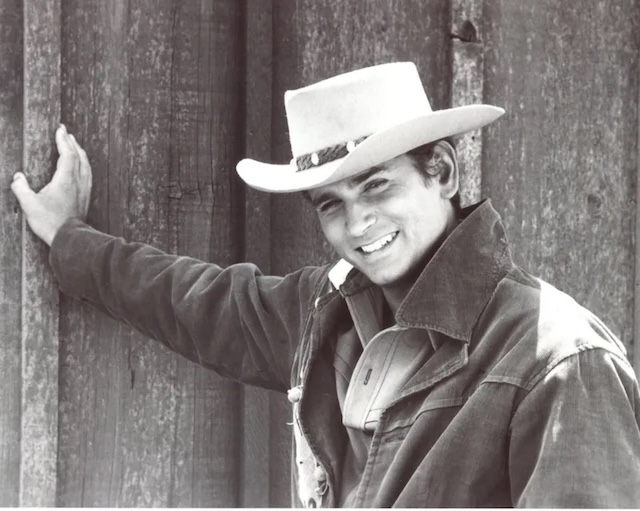 A close-up black-and-white portrait of a young Joe Cartwright, played by Michael Landon, leaning against a wall. His smile and youthful energy reflect the spirit of his character