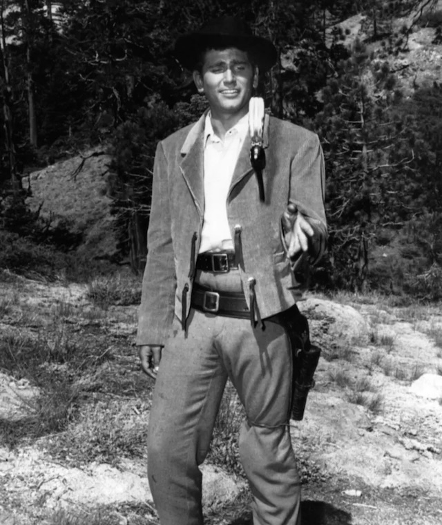 Joe Cartwright in a rugged outdoor scene, holding a pistol. His serious expression and stance are typical of his role as a cowboy defending his family and ranch