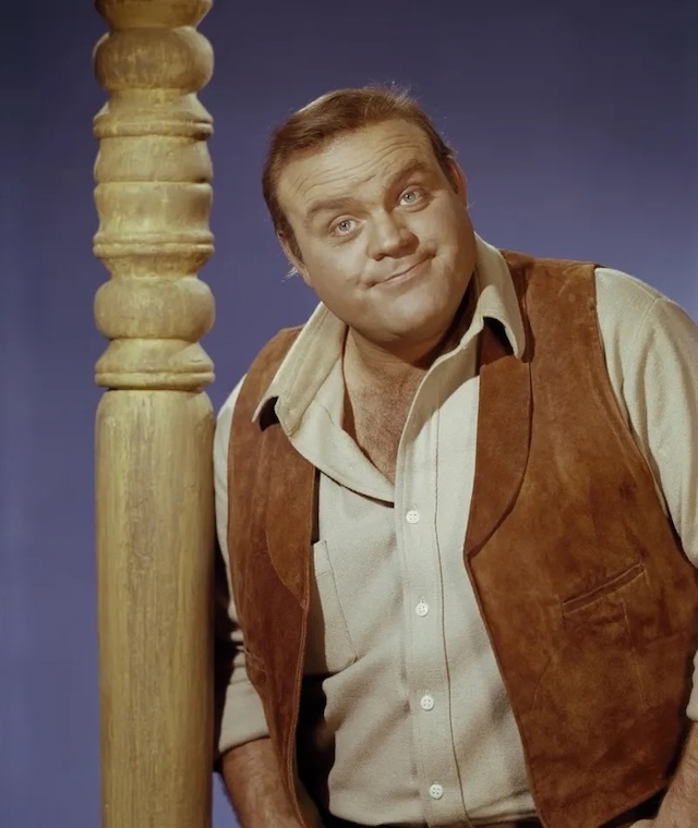 Another portrait of Hoss Cartwright, this time in color. He stands next to a wooden post, exuding his usual friendly and approachable nature that made him a fan favorite on Bonanza