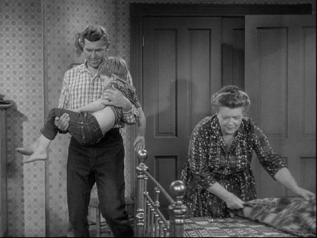 Andy Taylor gently carries a sleepy Opie while Aunt Bee makes the bed, showcasing the loving family dynamic that defined the series