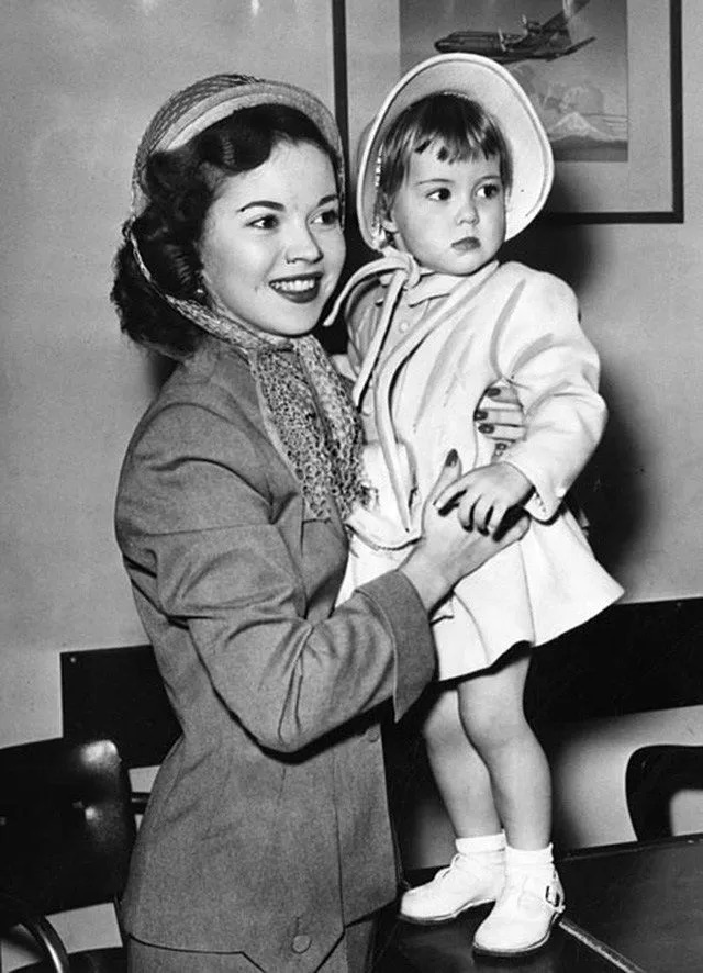 Dressed elegantly, Shirley Temple holds young Linda close while traveling, showcasing the protective and loving nature of their mother-daughter relationship.