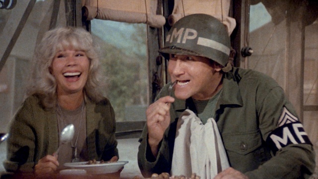 A candid scene from M*A*S*H featuring Loretta Swit and another character, sharing a laugh during a meal, reflecting the comedic elements of the show despite its war setting