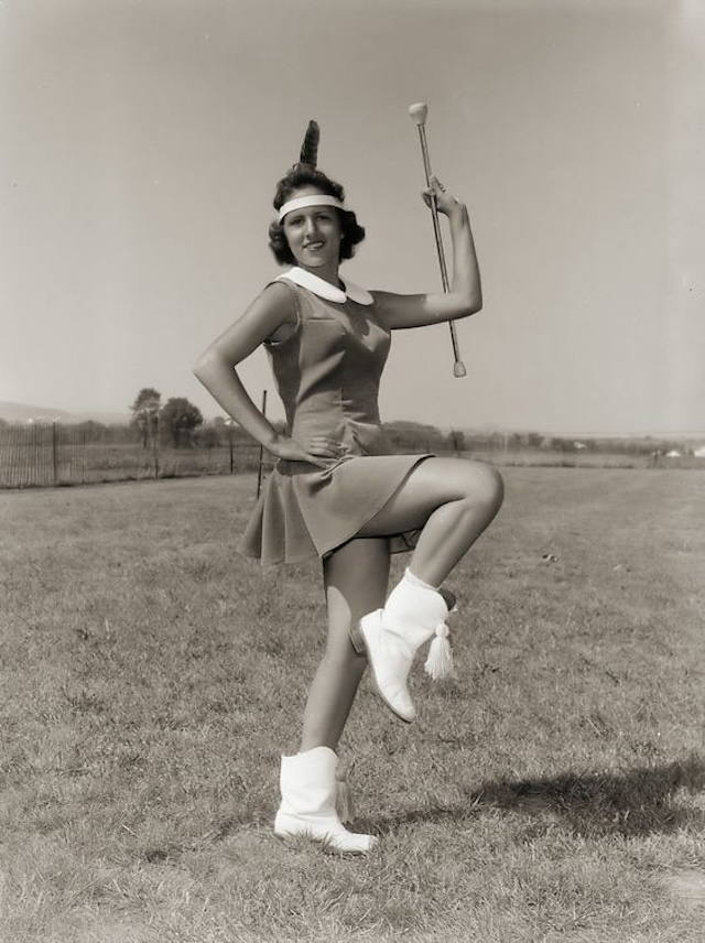 A vintage photo of a majorette proudly posing with her twirling baton in full costume, ready to lead a parade. This image captures the golden era of marching bands and baton twirling in the 1950s