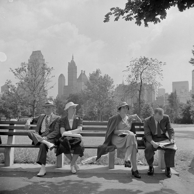 Four individuals sitting on a park bench, enjoying a quiet moment in Central Park, New York City, with the iconic skyline in the background. A snapshot of leisure in a bustling metropolis during the mid-20th century