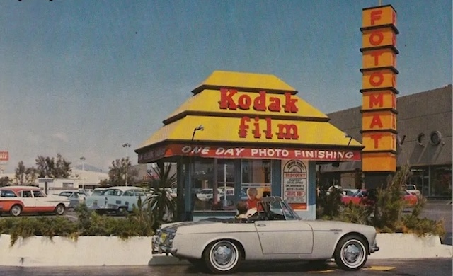 A customer pulls up to a Fotomat booth, part of mid-century suburban life, to drop off film for developing