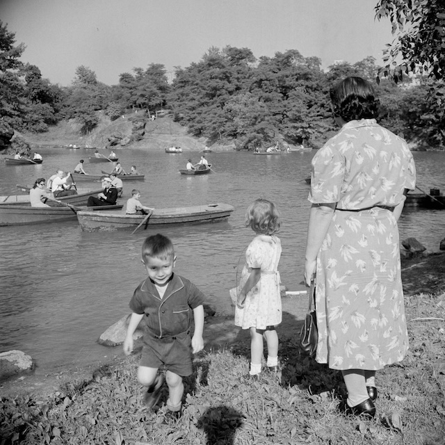 A mother watches her two children by the lakeside as rowboats glide across the water in Central Park. A typical family outing from decades ago, where spending time outdoors was a cherished tradition