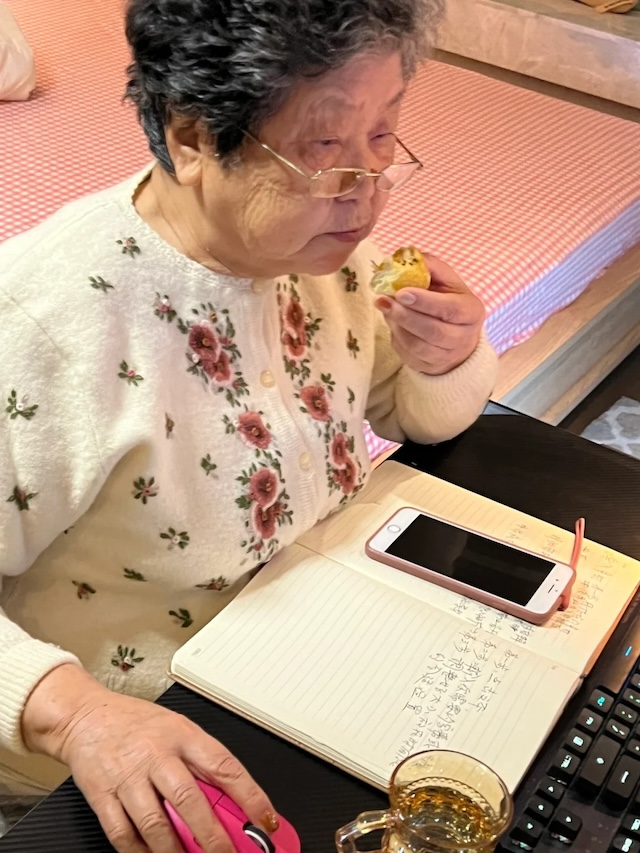 Taking a break while continuing to focus on her tasks, this determined grandmother enjoys a snack as she works through her video editing lessons