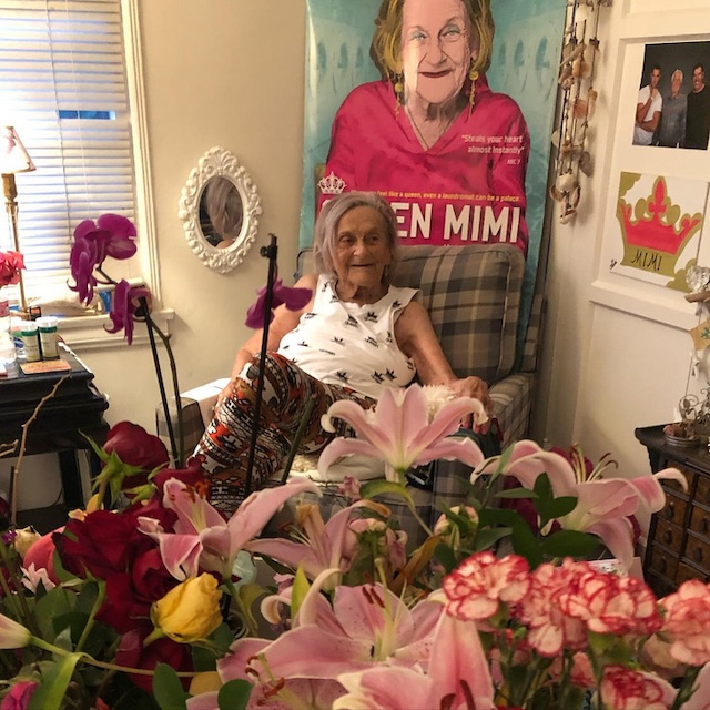 Surrounded by flowers, Mimi enjoys the warmth of her home, a far cry from the days of living on the streets. Her resilience and strength shine through as she sits in front of her "Queen Mimi" poster