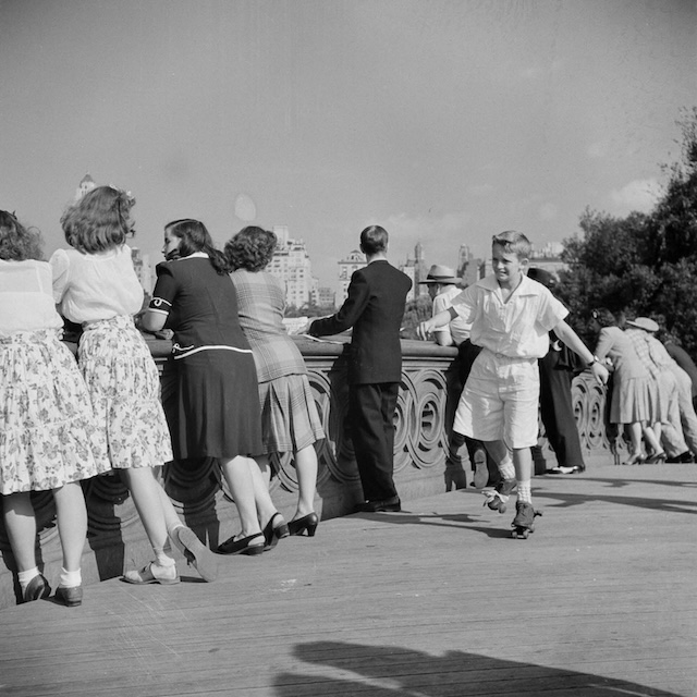 A young boy on roller skates speeds along the bridge in Central Park while others lean over to take in the view