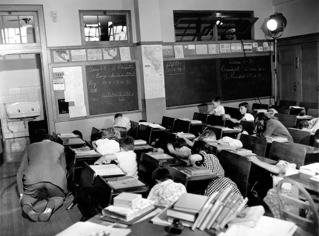 A 1950s civil defense drill, where children take cover under desks, a symbol of Cold War fears