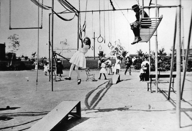 A girl gracefully navigates hanging rings while a boy perches atop the climbing structure. Playgrounds of the past had minimal supervision, letting children’s creativity lead the way