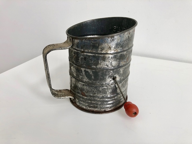 This vintage flour sifter features a red knob handle, adding a pop of color to its rustic metal body