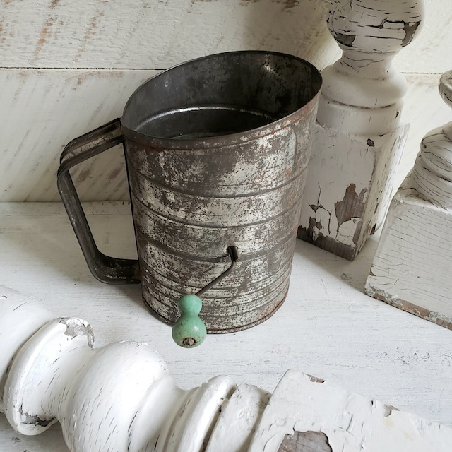 A beautifully aged flour sifter, complete with a green knob, placed decoratively among vintage home furnishings