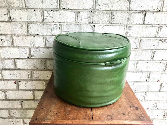 A beautifully preserved vintage green ottoman pouffe sitting gracefully against a brick wall. Its rich green color and smooth texture add an old-world charm to any home