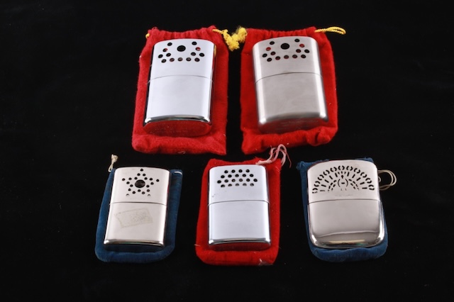 A collection of vintage hand warmers, including models with ornate designs, all nestled in their signature red and blue pouches