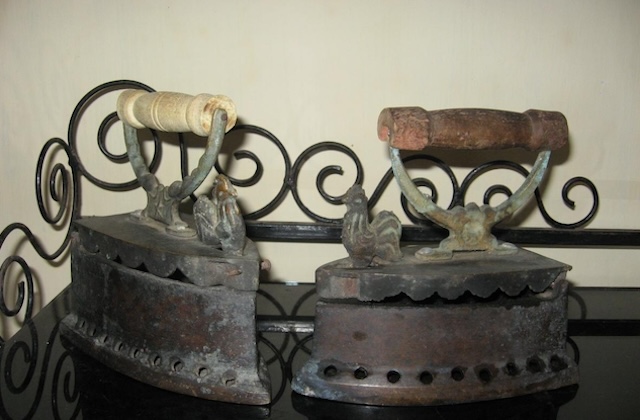 A pair of rustic antique coal irons with roosters on top – symbols of durability and craftsmanship. These irons were once used daily to press garments, their heavy metal build making them highly effective