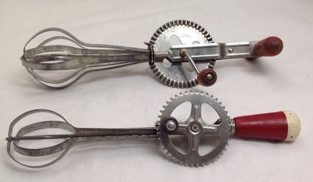 A pair of vintage hand-crank egg beaters, both featuring the iconic red wooden handles, a popular kitchen staple from the mid-20th century