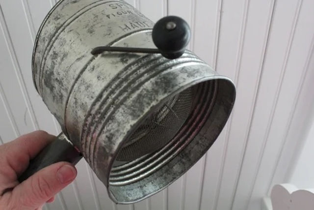 An overhead view of a classic flour sifter, still intact after years of use, held up for a closer look at its weathered details