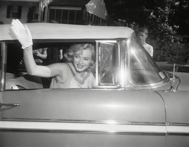 Marilyn Monroe waving cheerfully from the driver's seat, embodying her vibrant charm and love for classic cars