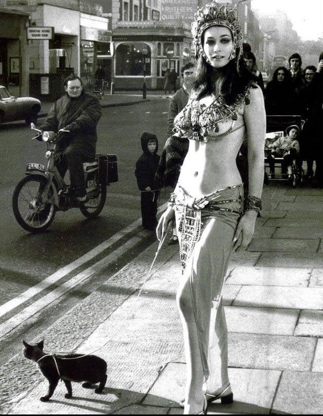 Valerie Leon struts through the streets in full Egyptian regalia, channeling the mystery and glamor of her iconic Mummy role, while passersby continue their everyday routines in the background