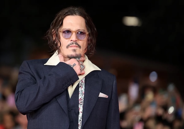 Depp poses on the red carpet, exuding his signature style with tinted glasses and an artistic scarf. His expression is both cool and contemplative, capturing the attention of fans and media alike