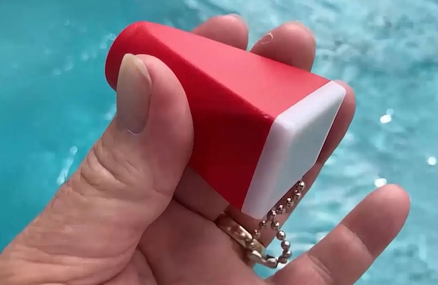 A hand gripping the red Vintage Delmar Colorscope picture viewer keychain, with a sparkling pool in the background, showcasing its compact and nostalgic design