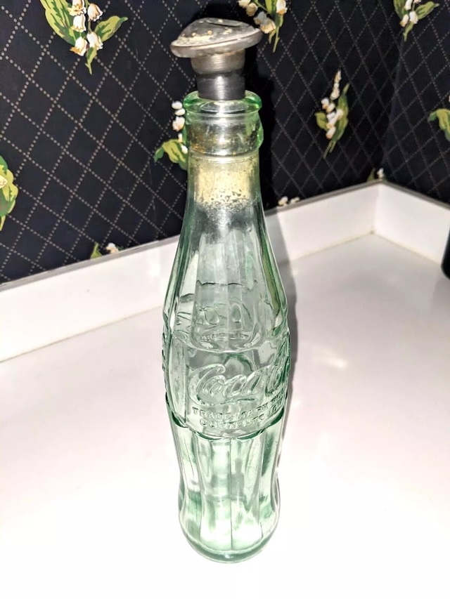 The vintage glass sprinkler bottle standing upright, featuring a green-tinted glass body and a metal sprinkler head attached