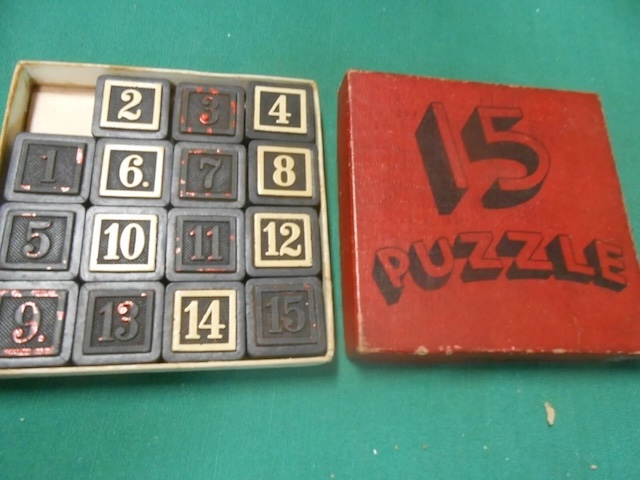 A vintage boxed 15 Puzzle set featuring black and white tiles with bold numbers, paired with a striking red cover emblazoned with "15 Puzzle."