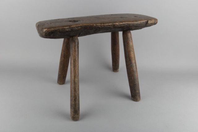 A side profile of an individual traditional wooden milking stool, emphasizing its simple yet effective three-legged design