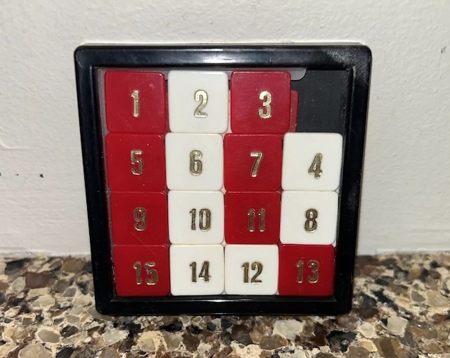 A nostalgic 15 Puzzle with alternating red and white tiles housed in a black frame, evoking memories of hours spent solving its engaging challenge