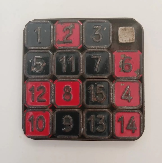 A well-worn 15 Puzzle with aged red and black tiles, bearing marks of countless hours of play, testifying to its enduring appeal