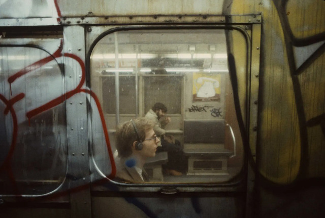 A man, with headphones on, gazes through the subway window, losing himself in music amidst the urban cacophony of 1981.