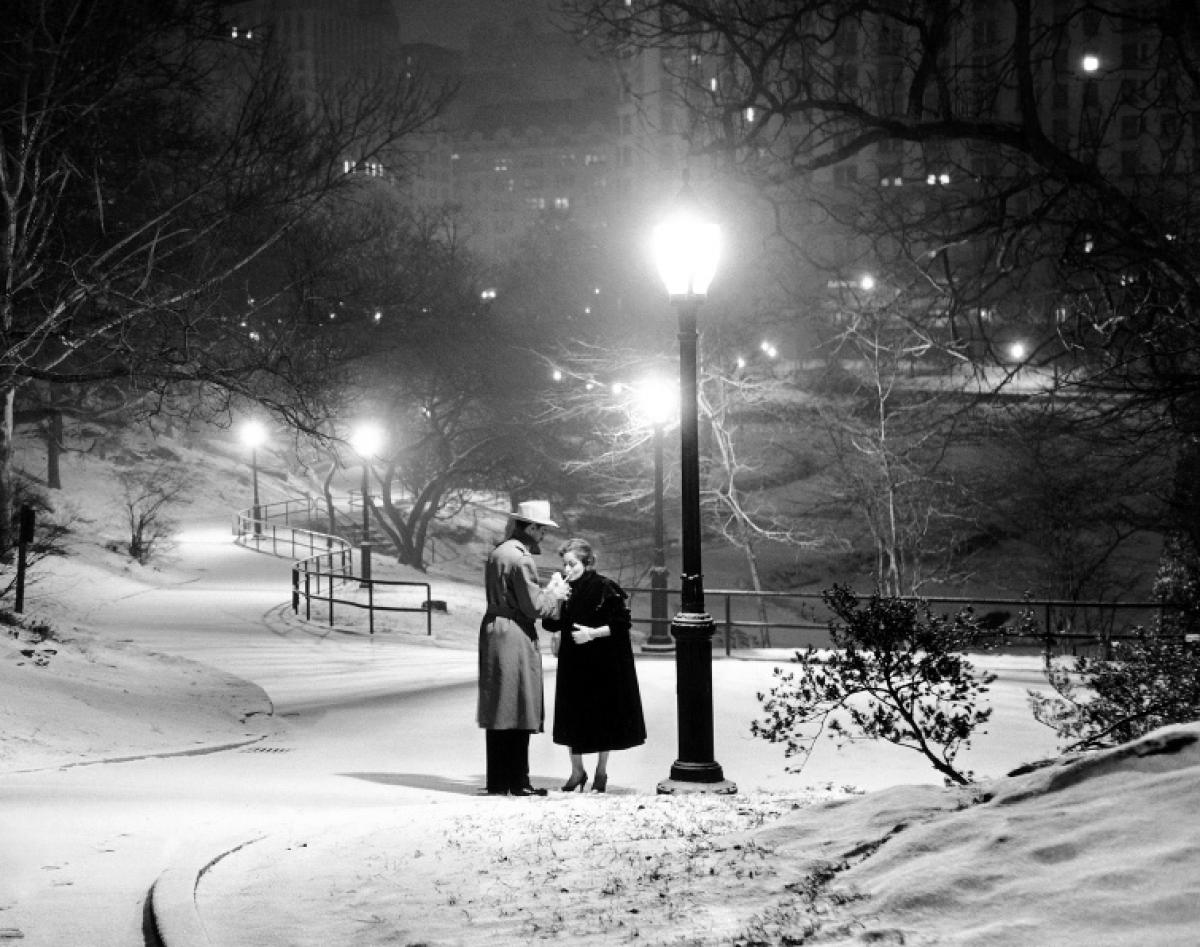 Against the glowing city lights, a couple shares a cigarette under a street lamp in Central Park, warding off the cold together, 1957.