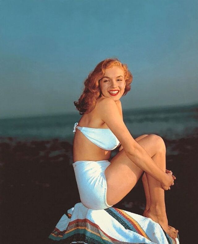 A sunlit seaside photo of Norma Jeane sitting on a rock in a white swimsuit, capturing the carefree spirit of her earliest modeling days.